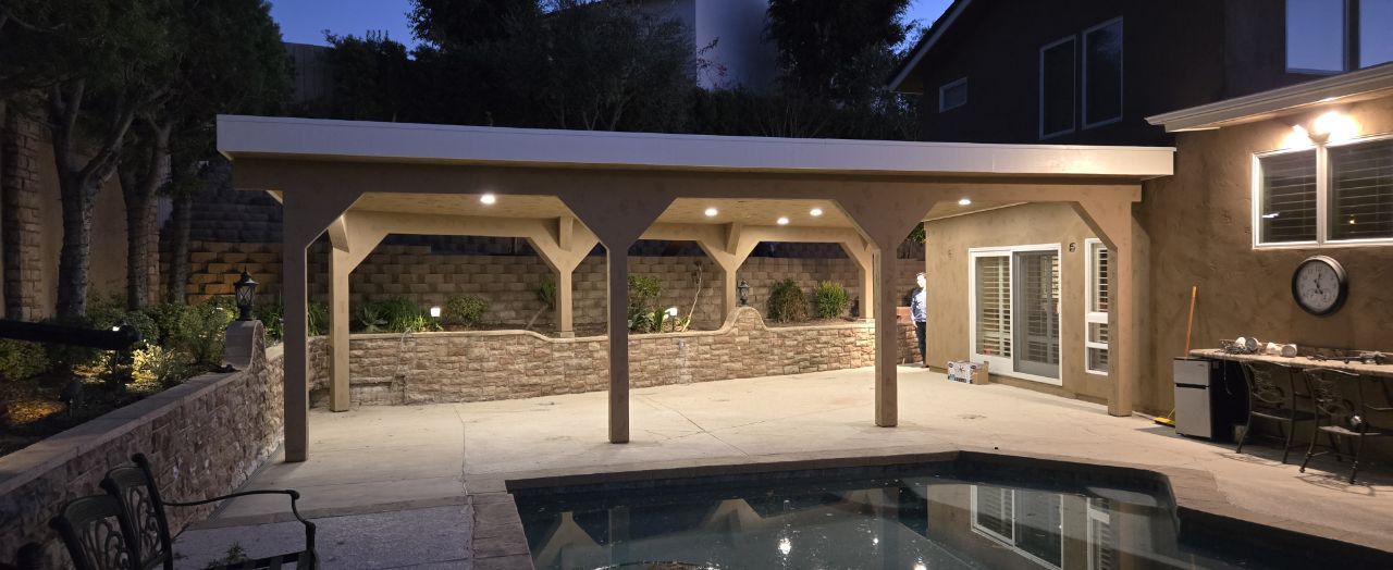 A light-colored wooden patio cover beside a backyard pool, illuminated by recessed ceiling lights at night.