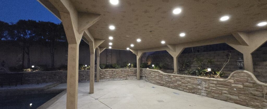 A wide covered patio at dusk with multiple recessed ceiling lights illuminating the stone planter wall and open walkway.
