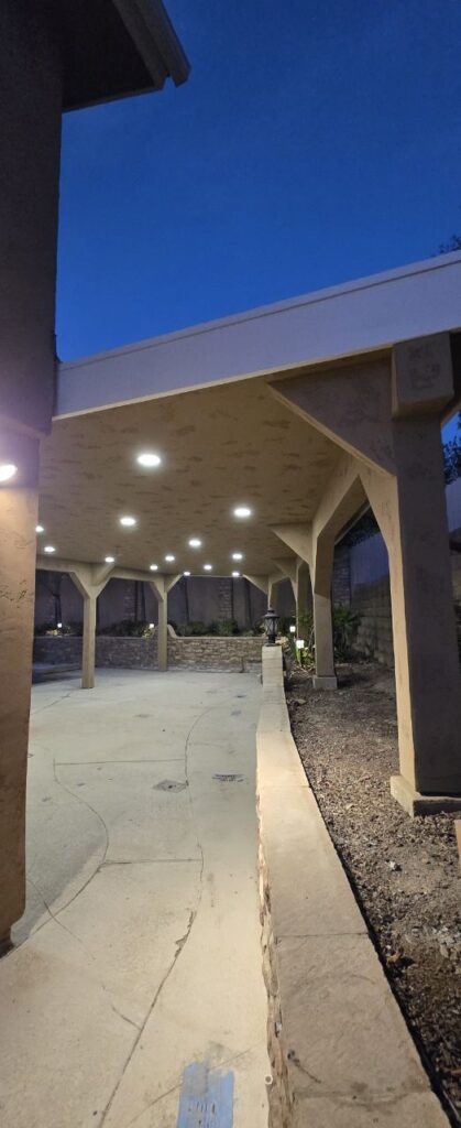 A covered patio structure at dusk with multiple recessed lights illuminating the walkway and stone retaining wall.