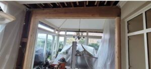 Interior renovation scene showing an exposed ceiling, newly installed wooden beam, and construction materials covered with plastic sheeting